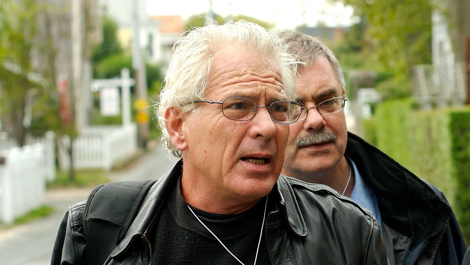 Chris Busa, white-haired, in glasses and a leather jacket, talking as he walks a Provincetown street